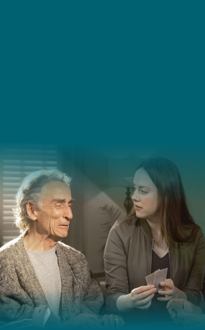 Actor portrayals of an older male experiencing Pseudobulbar Affect (PBA) crying symptoms, next to a younger female holding playing cards