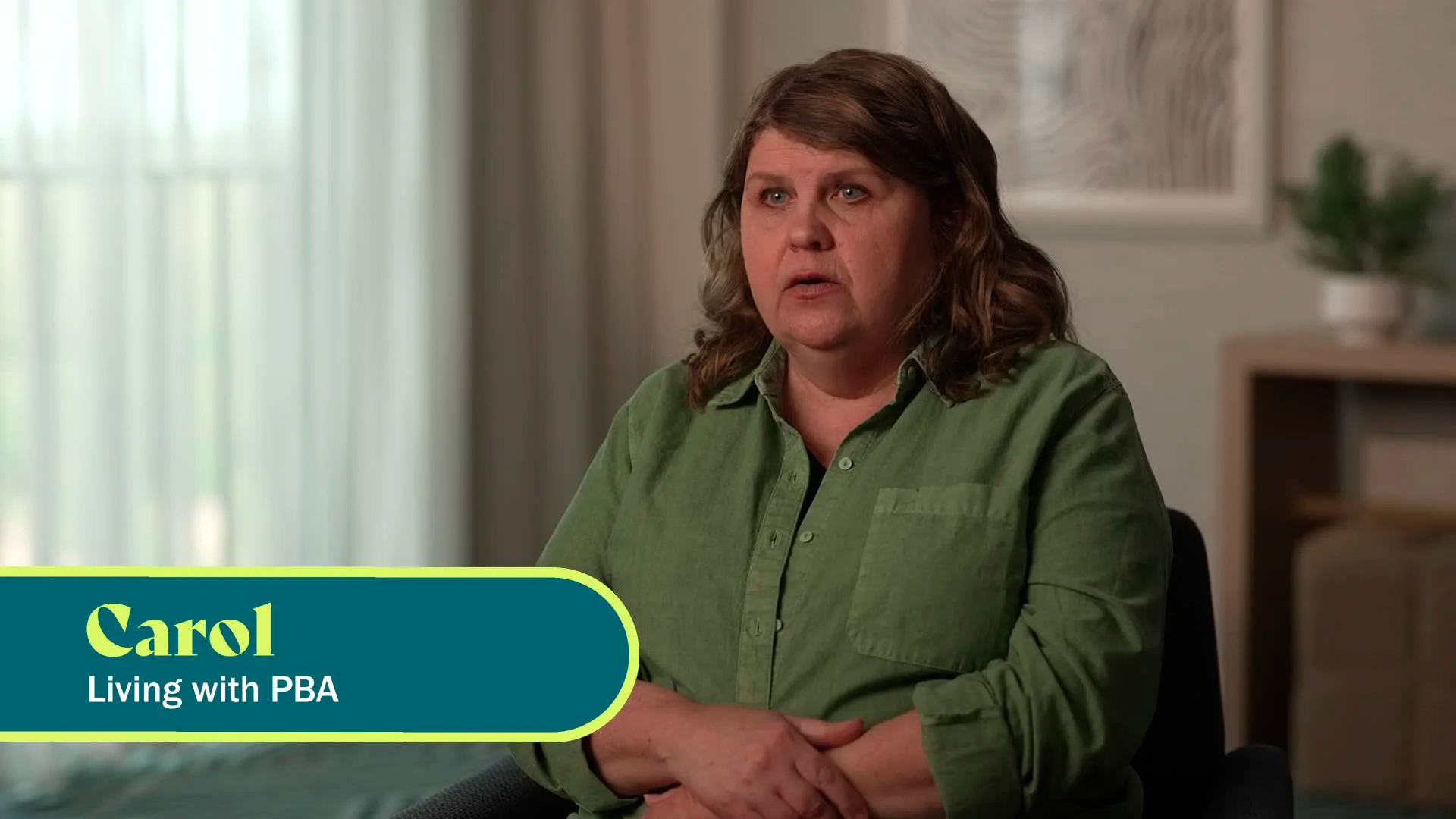 A white woman with wavy brunette hair is seen from the waist up, facing the camera. The caption on screen says “Carol, living with PBA.”