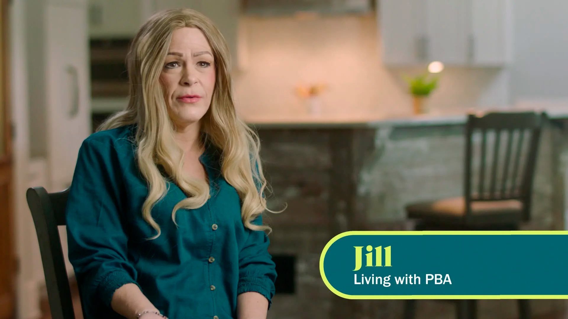 A white woman with long blonde hair is seen from the waist up, facing the camera. The caption on screen says “Jill, living with PBA.”